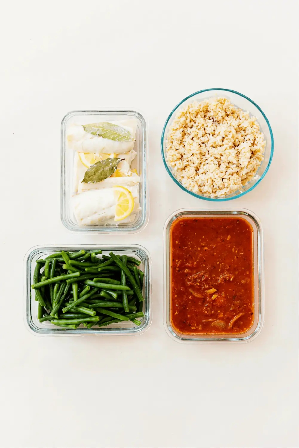 Glass dishes filled with fish, rice, sauce and green beans. Each item is in a separate container. The photo is from an overhead view with a white background.