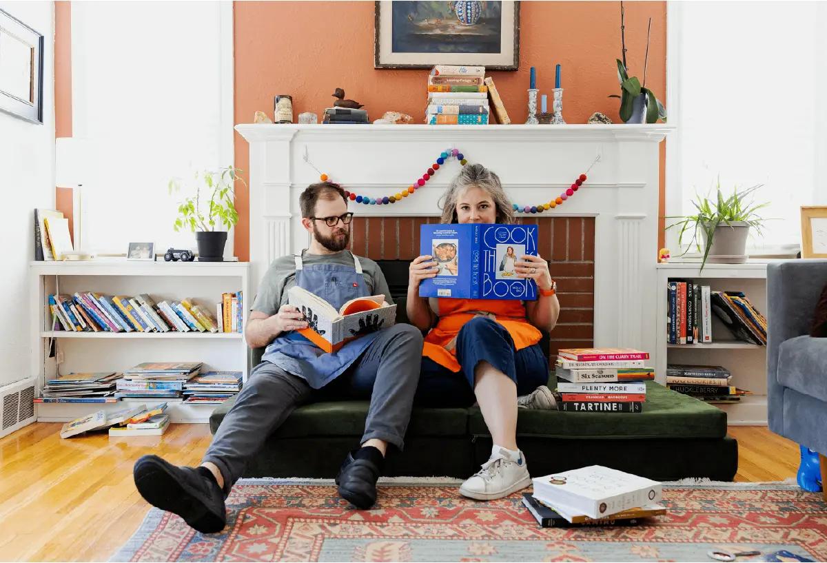 A married couple wearing aprons and reading cookbooks while sitting on couch cushions